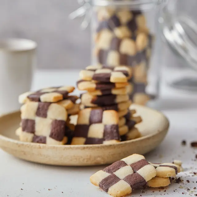 ⁨	עוגיות שחמט וניל ושוקולד יפהפיות על השיש בצנצנת ומעולות בימים קרירים לצד התה💕 
איך מכינים? 
מצרכים:
200 גרם חמאה קרה, חתוכה לקוביות
100 גרם אבקת סוכר
250 גרם קמח
1 ביצה M
2 כפות מים - במידת הצורך
4 כפות שוקולית
3 כפות חלב

אופן ההכנה:
- במעבד מזון שמים חמאה, אבקת סוכר וקמח ומעבדים בפולסים עד לקבלת תערובת שמזכירה בורגול.
- מוסיפים ביצה וממשיכים לעבד עד שנוצר בצק שמתגבש סביב וו המכשיר – אם הבצק יבש אפשר להוסיף 2-3 כפות מים. למי שאין מעבד מזון בבית ניתן להכין את הבצק עם מיקסר ו-וו גיטרה.
- מוציאים מחצית מהבצק החוצה, מעבדים עם הידיים למעין פיתה ועוטפים בניילון. מעבירים למקרר לחצי שעה.
- ליתרת הבצק במעבד מזון/מיקסר מוסיפים שוקולית וחלב ומעבדים עד שהבצק נצבע בצבע שוקולדי חלק.
- מעבדים מעט עם הידיים, מועכים למעין פיתה ושומרים במקרר חצי שעה עטוף בניילון.
- מרדדים כל אחד מהבצקים לעלה בעובי 1 ס”מ (ולא פחות מכך) וחותכים לרצועות בעובי 1 ס”מ. בעצם מה שיווצר לכם זה רצועות בצק בעובי וברוחב של 1 ס”מ. מומלץ שכל הרצועות תהיינה באותו אורך.
- מניחים 3 רצועות אחת ליד השנייה ומוודאים שהן נצמדות ונדבקות אחת לשנייה. קודם רצועה לבנה, אחר כך כהה ולאחר מכן שוב לבנה.
- מעל מניחים שוב 3 רצועות בצק הפעם קודם כהה אחר כך לבן ולבסוף כהה. בשורה מעל חוזרים על סדר של רצועה בהירה, כהה ולבסוף בהירה. שימו לב – אם רצועות הבצק לא נצמדות לכם ניתן להבריש את החיבור ביניהן עם חלבון.
- מעבירים את המרובע עם 9 הרצועות בתוכו, בזהירות, למקפיא לכ-שעה (מומלץ לעטוף עם נייר אפייה). אם נשארו רצועות בצק נוספות מכינים גם איתן מלבן ארוך עם 9 רצועות.
- מחממים תנור ל-180 מעלות.
- פורסים את פס הבצק המחולק לפרוסות בעובי 1/2 ס”מ ומסדרים על תבנית תנור עם נייר אפייה.
- אופים במשך 15 דקות או עד הזהבה עדינה ומצננים. שומרים בצנצנת בחוץ.⁩