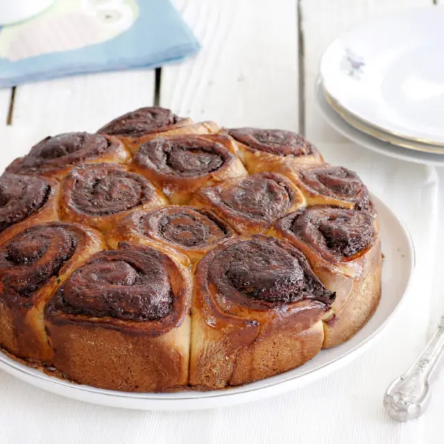 שילוב משגע של קפה ושוקולד בתוך עוגת שבלולי שמרים. בצק נוח לעבודה, ניחוח מתוק שקשה לעמוד בפניו ורגע אחד מתוק בבוקר שבת💕
איך מכינים?
לבצק:
500 גרם (3 וחצי כוסות) קמח
10 גרם (1 כף) שמרים יבשים או 30 גרם שמרים טריים
100 גרם (1/2 כוס) סוכר
125 גרם חמאה רכה
200 מ”ל (1 מכל) שמנת חמוצה
1 ביצה L
קורט מלח
1 כפית תמצית וניל

למלית שוקולד וקפה:
200 גרם שוקולד פרה מריר
100 גרם חמאה
2 כפיות קפה נמס

לסירופ קפה:
80 מ”ל (1/3 כוס) מים
70 גרם (1/3 כוס) סוכר
1 כפית קפה נמס

להברשה:
ביצה טרופה

אופן ההכנה:
- בצק: בקערת מיקסר שמים קמח, שמרים, סוכר, חמאה, שמנת חמוצה, ביצה, מלח ווניל ולשים במהירות נמוכה-בינונית במשך 10-12 דקות או עד שמתקבל בצק אחיד וחלק.
- מכסים את הקערה ומתפיחים את הבצק במשך כשעה-שעתיים בטמפרטורה חמימה או עד שהוא מכפיל את נפחו.
- מלית שוקולד וקפה: שוברים את השוקולד לקוביות ושמים בקערה. מוסיפים חמאה וקפה וממסים יחד במיקרוגל או על בן מארי עד שהכל נמס והתערובת אחידה.
- מצננים את התערובת במקרר עד שהיא מגיעה למרקם של ממרח.
- הרכבת העוגה: מחלקים את הבצק ל-2 חלקים.
על משטח מקומח מרדדים כל חלק למלבן בעובי 3-4 מ”מ ויוצקים מחצית מכמות המלית. מורחים בשכבה אחידה ודי דקה ומגלגלים לרולדה הדוקה.
- חותכים את הרולדה לחתיכות באורך 5-6 ס”מ (שושנים) ומניחים במרווחים של 1-2 ס”מ זו מזו בתוך התבנית המשומנת.
- חוזרים על הפעולה עם החלק השני של הבצק ויתרת המילוי ליצירת שושנים נוספות.
- מכסים את העוגה ומתפיחים במשך 45-75 דקות או עד שהשושנים כמעט מכפילות את נפחן.
- מחממים תנור ל-170 מעלות.
- מברישים את העוגה התפוחה בביצה טרופה בעדינות.
אופים במשך 30-40 דקות או עד שהעוגה תפוחה, יציבה והשושנים זהובות.
- סירופ קפה: בסיר קטן שמים מים, סוכר וקפה ומבשלים יחד על להבה בינונית עד שהסוכר והקפה נמסים לחלוטין ומתקבל סירופ.
- מברישים את העוגה בסירופ ומניחים לה להצטנן בטמפרטורת החדר.